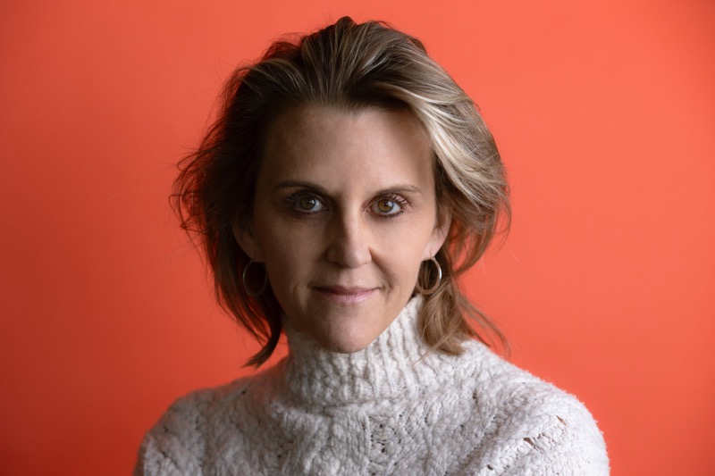 A person with shoulder-length blond hair is shown from the shoulders up against a solid orange background, wearing a light-colored cable-knit turtleneck sweater. The lighting is even, illuminating the face with soft shadows. The expression is neutral, with direct eye contact. The composition is centered and closely cropped.