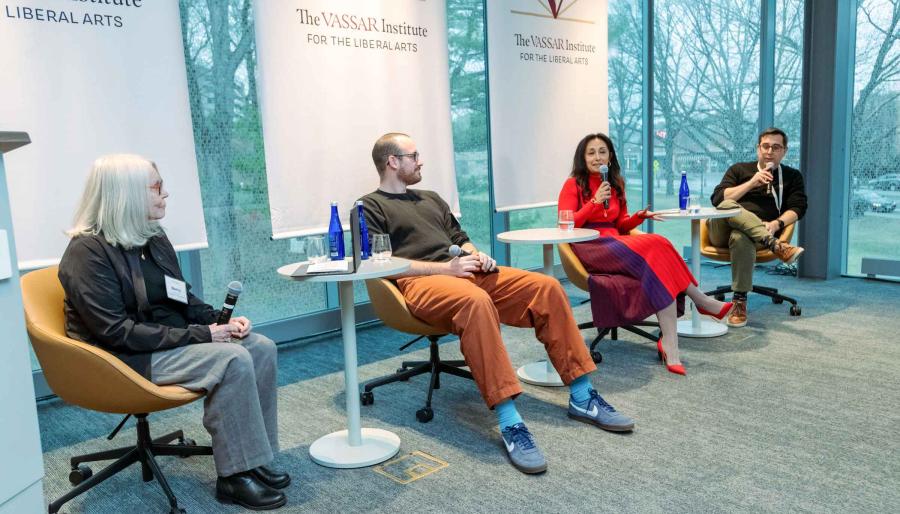 A seated panel of four people listen while one of the panelists holds and speaks into a microphone.