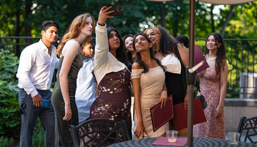 A group of young adults, dressed in elegant attire, gather under an outdoor umbrella for a selfie. 