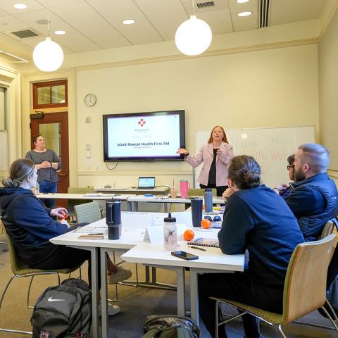 Four people in a classroom sitting at a desk/table looking at a speaker with a TV presentation in the background.